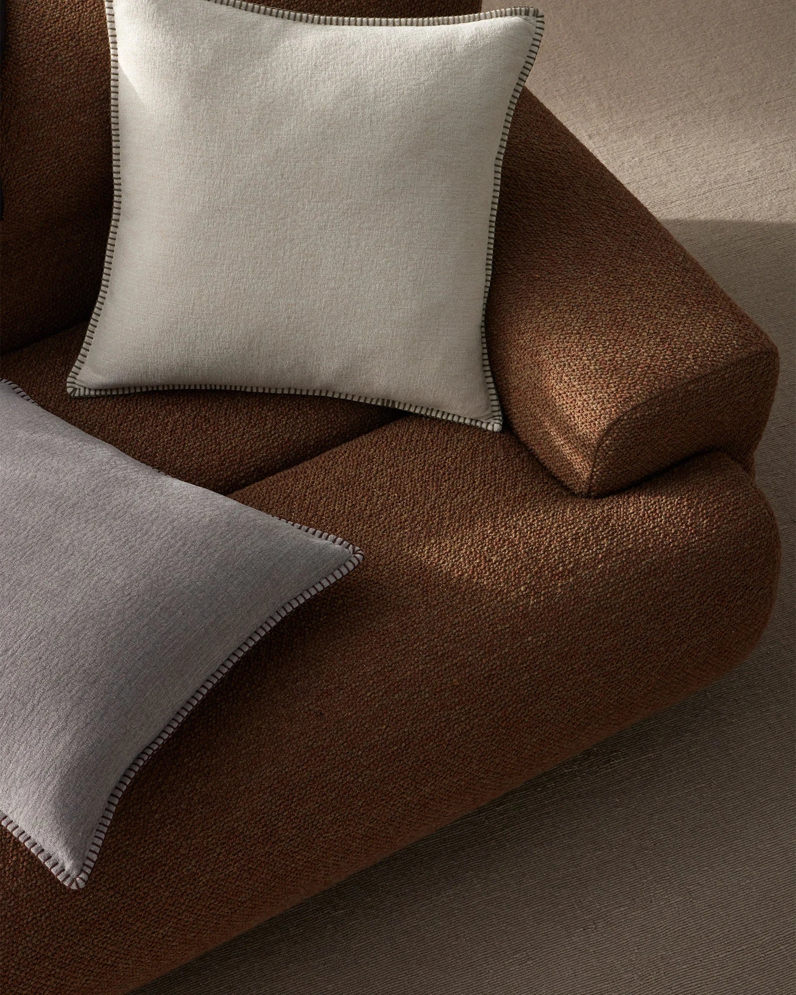 Neutral and chocolate brown textiles layered on a sofa, featuring Alba cushions with contrast blanket stitching in a warm, earthy palette with natural linen textures