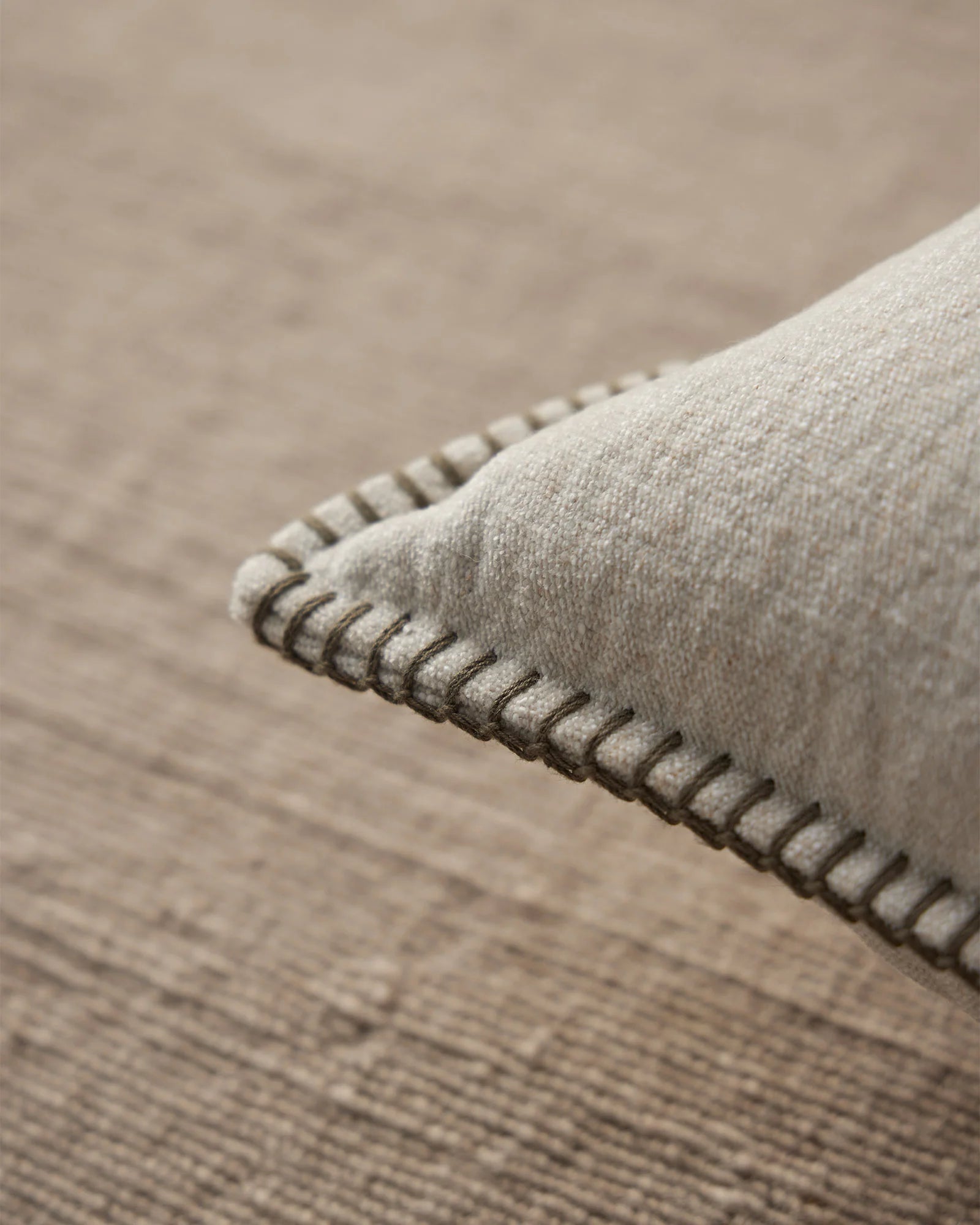 Neutral linen cushion with blanket stitch edging detail, draped on warm taupe textured fabric in monochromatic interior styling