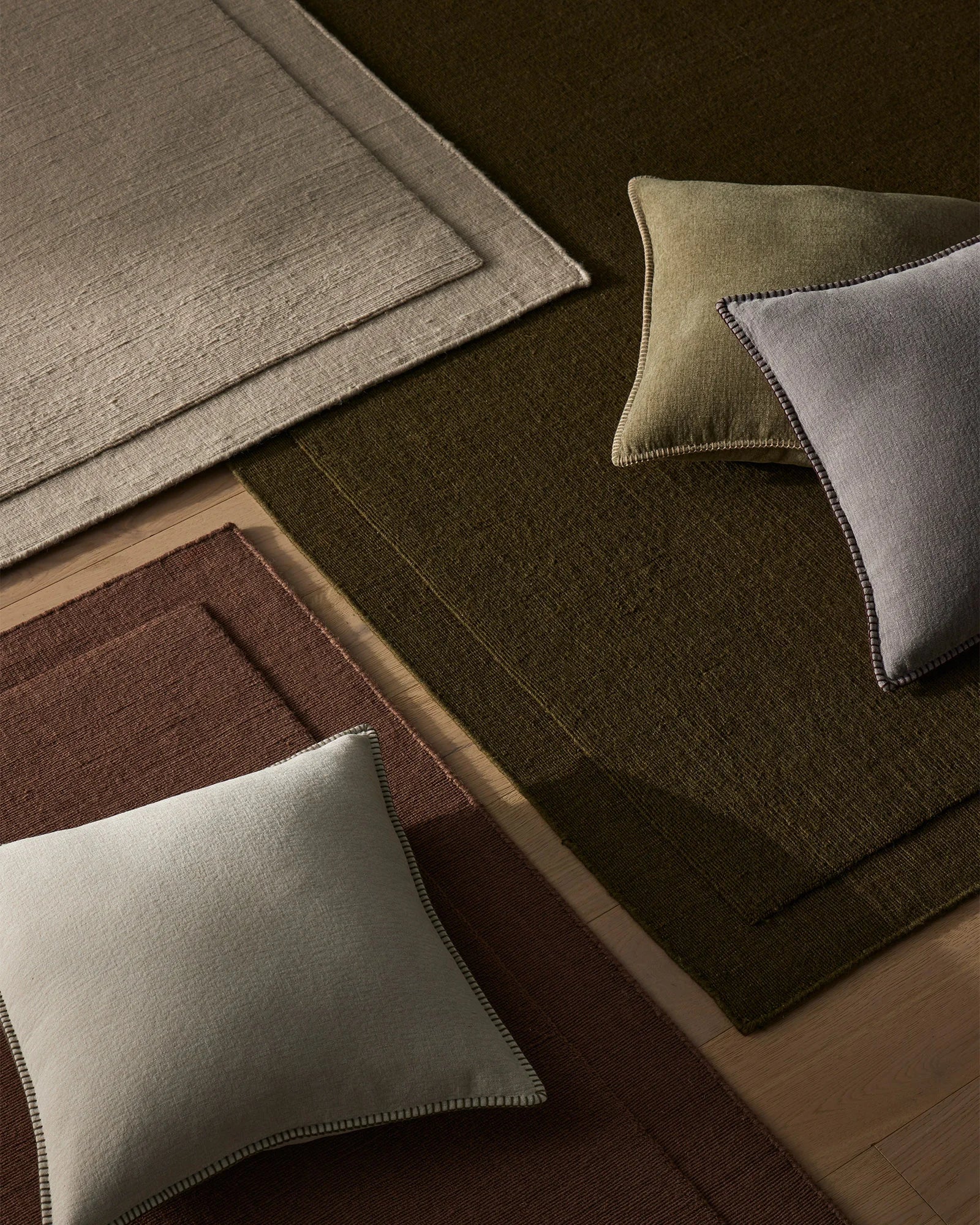 Earthy wool rugs in olive, mulberry and sage tones layered with linen cushions in bone, dove grey and moss, showcasing natural texture and warm neutral styling