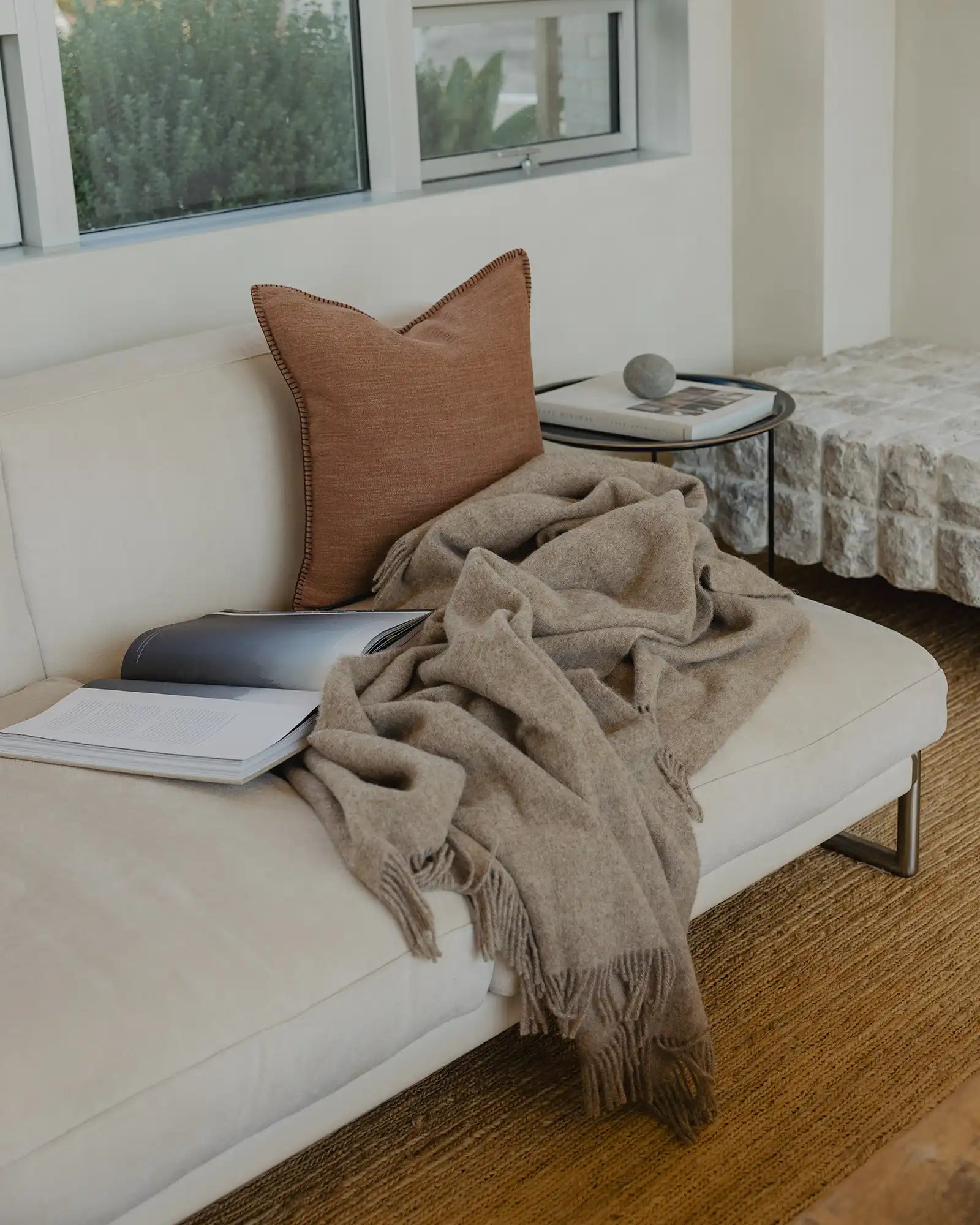 Minimalist workspace with warm earth tones: taupe wool throw draped over cream seating, rust linen cushion, and natural fibre rug against floor-to-ceiling windows with forest views