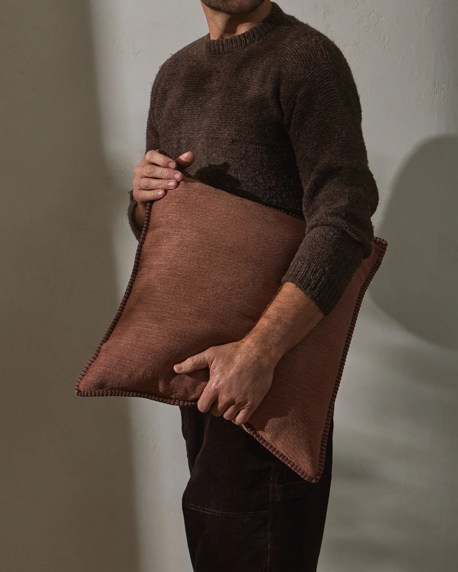 Man holding rust linen cushion against brown knit sweater, earthy tonal palette with textured fabrics and minimalist styling in natural light
