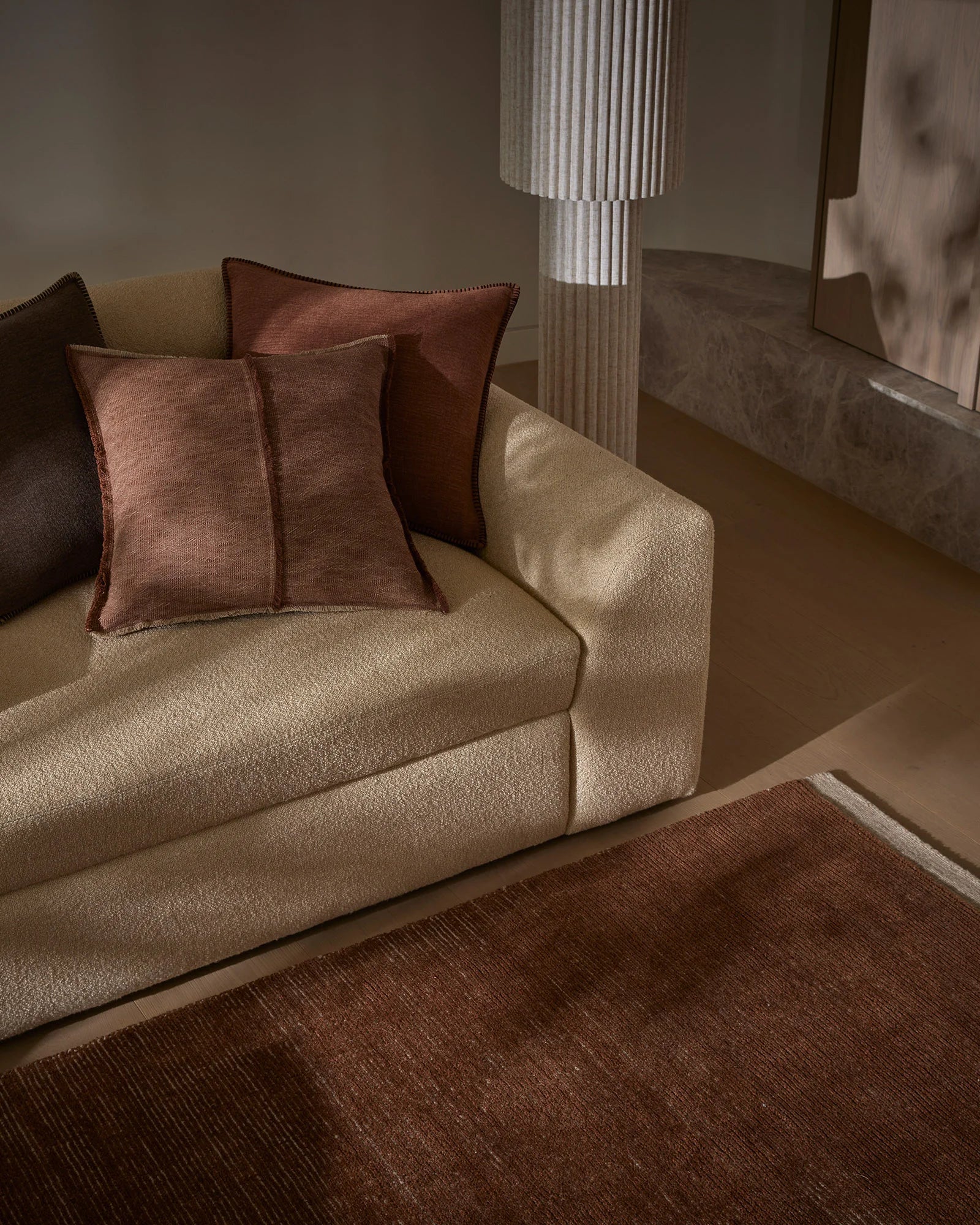 Warm earthy living room with cream sofa, rust and chocolate cushions, and deep brown wool rug under soft pendant lighting