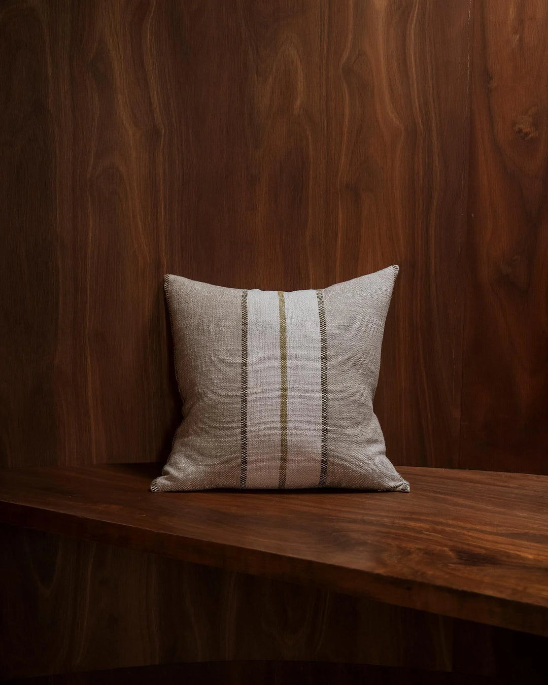 Neutral linen cushion with sage stripe detail on dark wood surface, warm timber backdrop. Earthy woven textile with minimalist styling and organic colour palette.