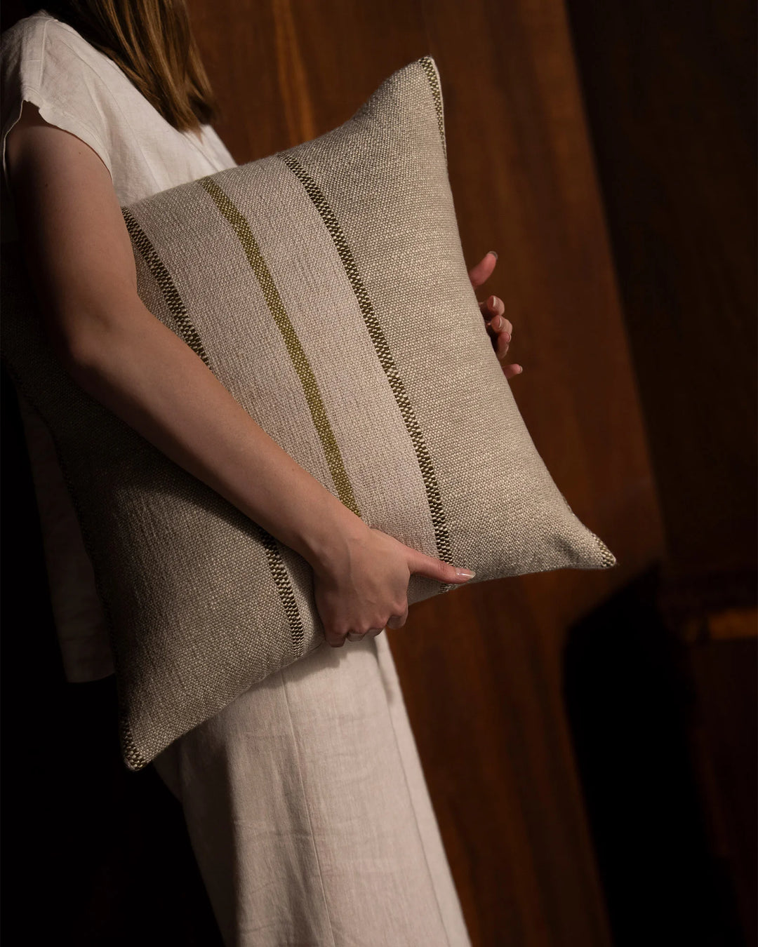 Neutral linen cushion with subtle vertical stitch detail held against warm ochre-toned drapery, styled with cream linen clothing for organic, minimal aesthetic