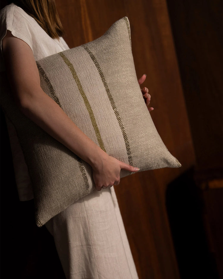 Neutral linen cushion with subtle vertical stitch detail held against warm ochre-toned drapery, styled with cream linen clothing for organic, minimal aesthetic