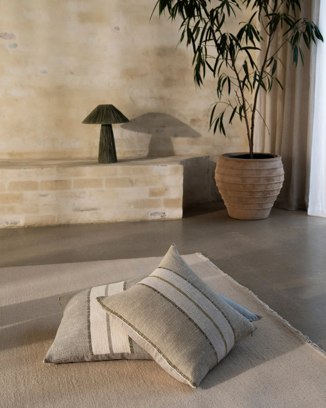 Neutral linen cushions with sage striping layered on cream carpet in minimalist room with brick wall, wooden planter and cascading foliage