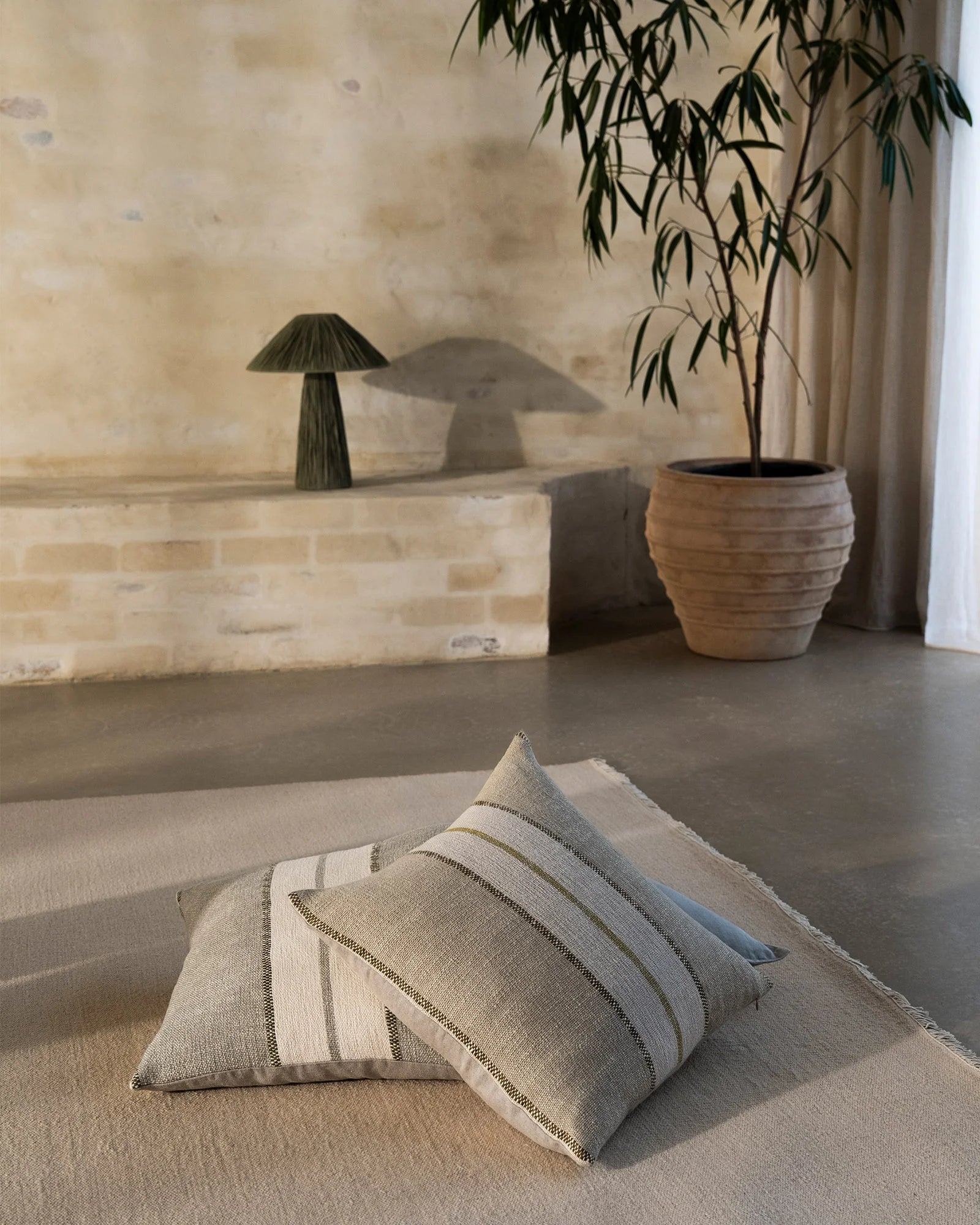 Neutral linen cushions with sage striping layered on cream carpet in minimalist room with brick wall, wooden planter and cascading foliage