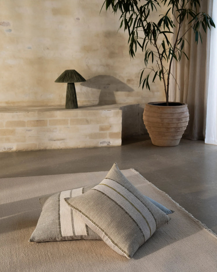 Neutral linen cushions with sage striping layered on cream carpet in minimalist room with brick wall, wooden planter and cascading foliage