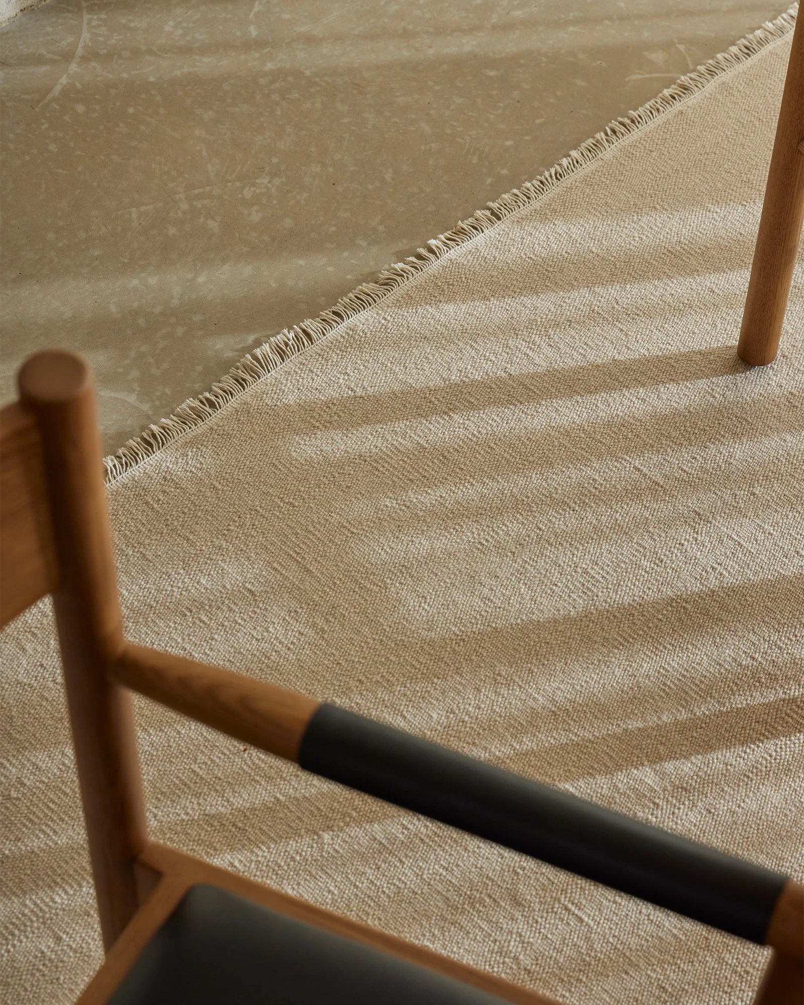 Neutral wool rug in warm cashew tone layered beneath mid-century timber furniture, showcasing textured weave and natural fringe detailing in minimalist interior styling