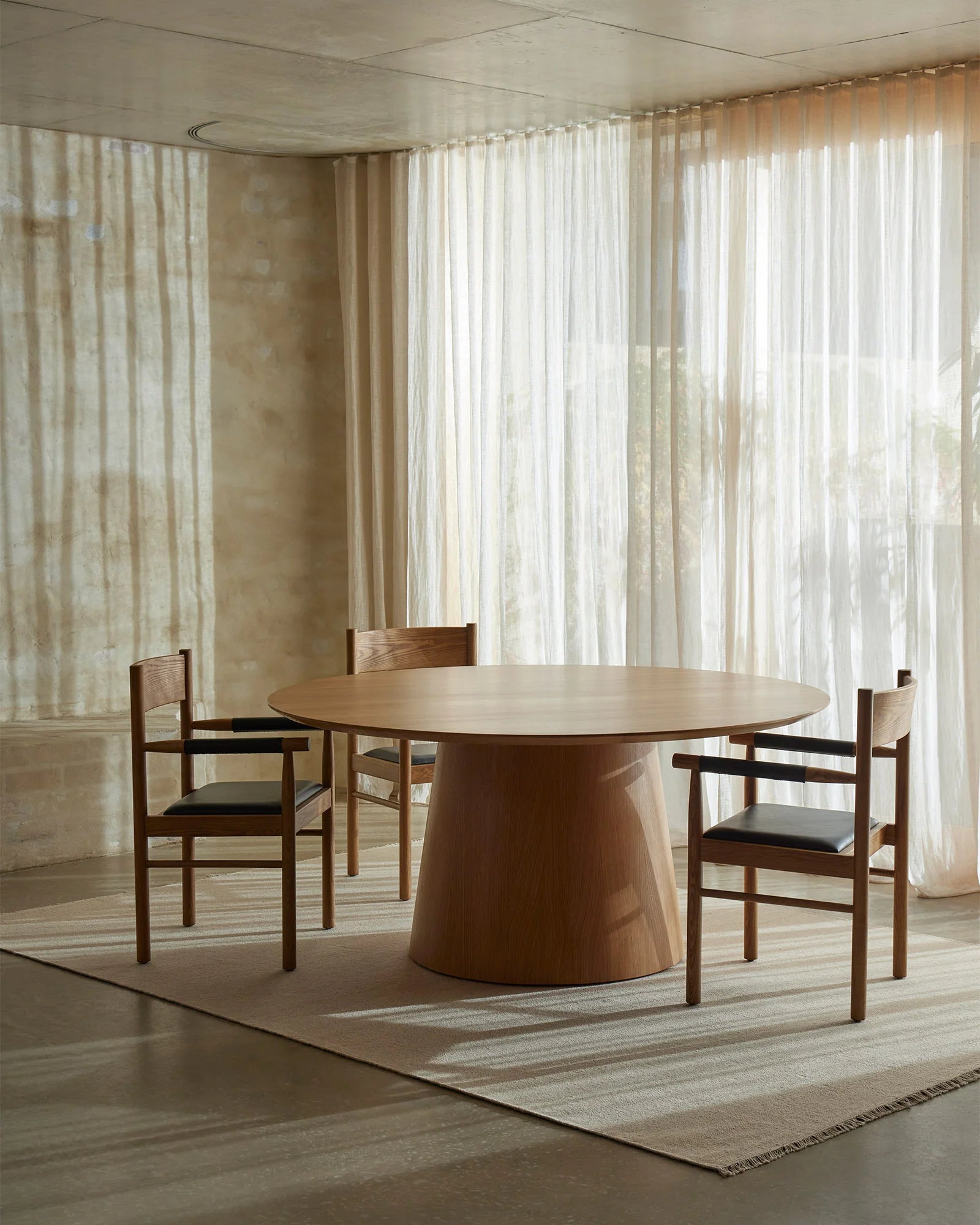 Warm neutral dining space with Jacqueline Cashew rug anchoring round timber table, sheer cream curtains filtering diffused light, mid-century modern aesthetic