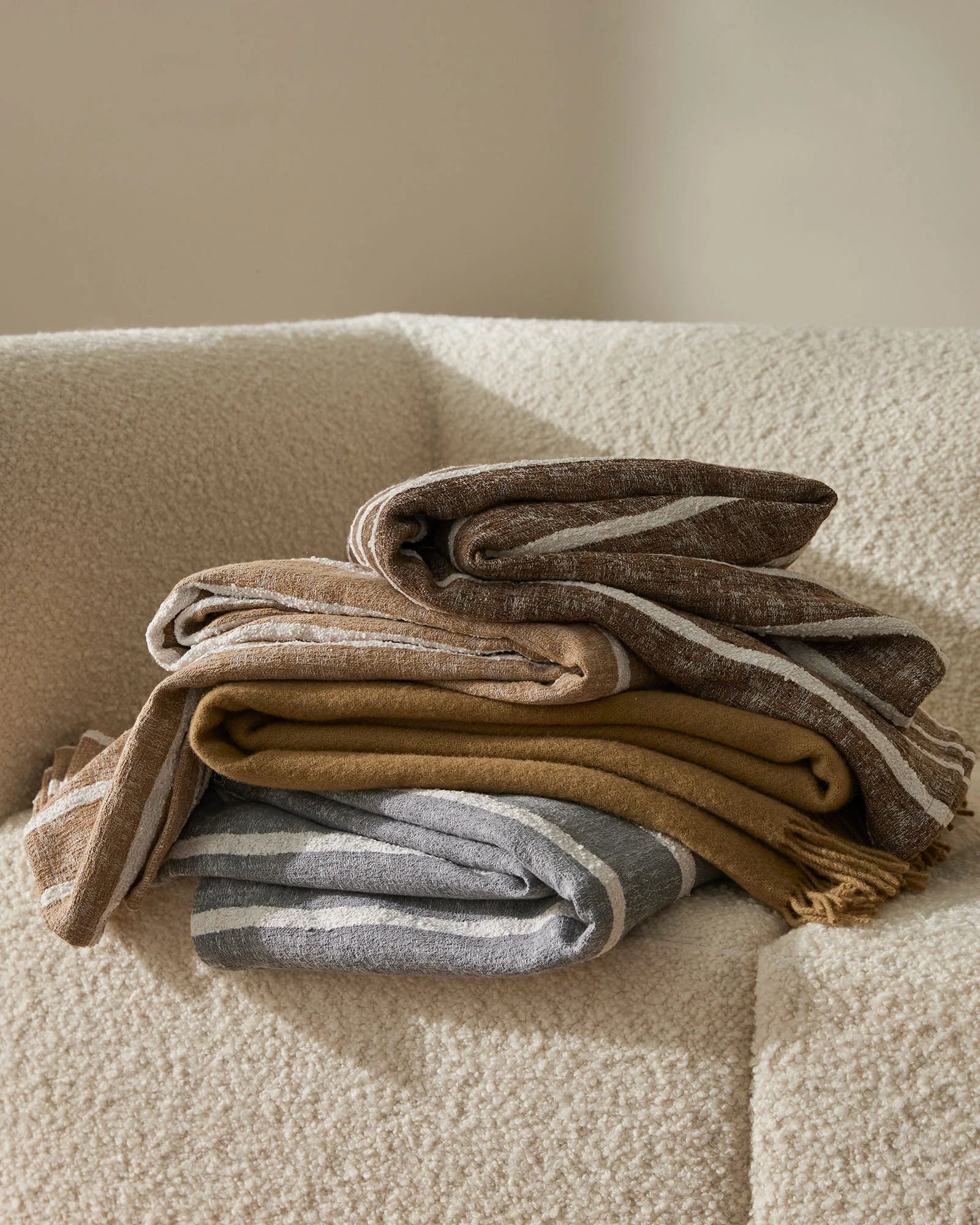Neutral earth tone throws stacked on cream linen sofa, layering warm taupe, ochre, and soft grey in textured cotton blends against minimal backdrop
