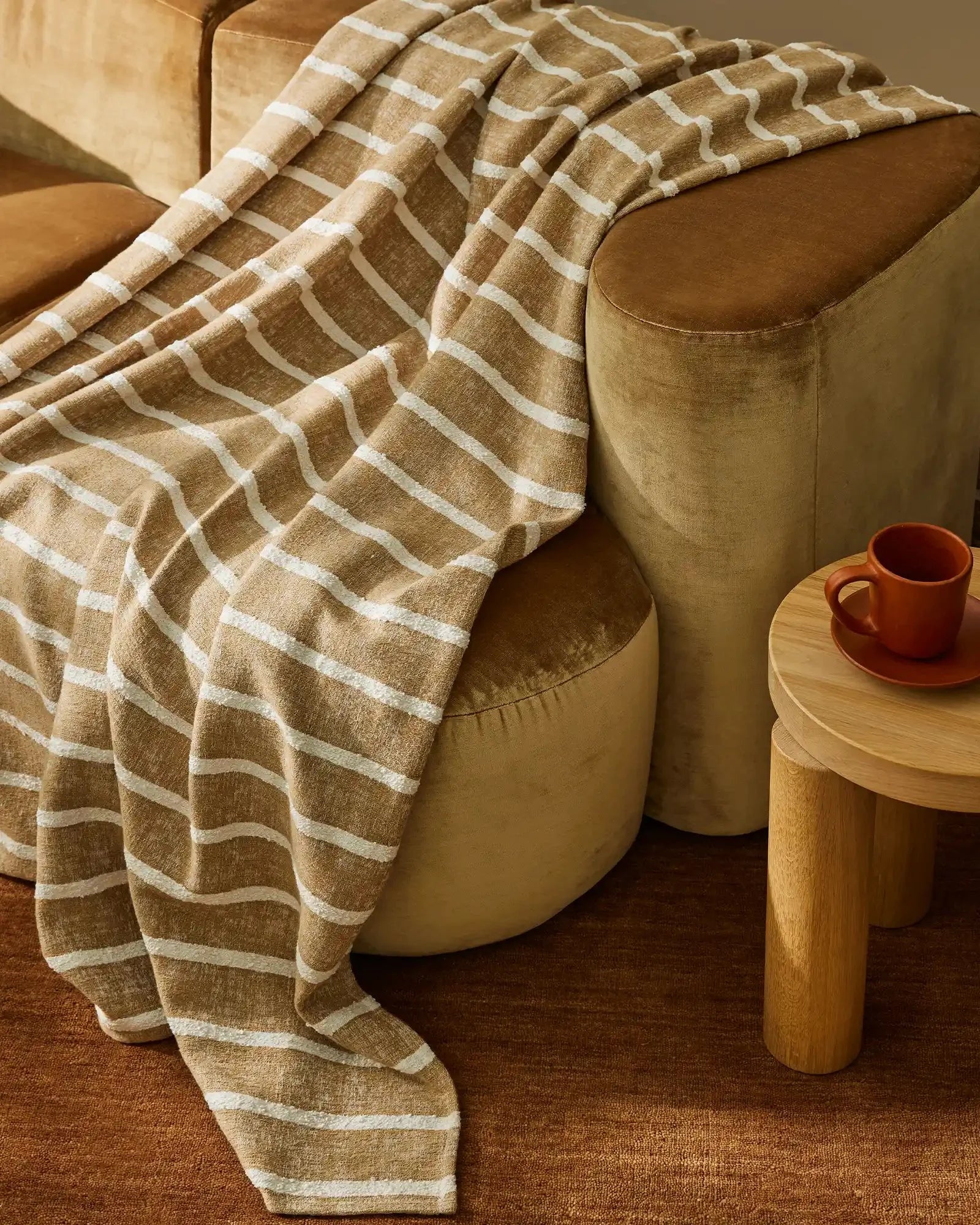 Warm neutral living room with tan linen sofa draped in geometric patterned throws in caramel and cream, styled with wooden side table and terracotta mug