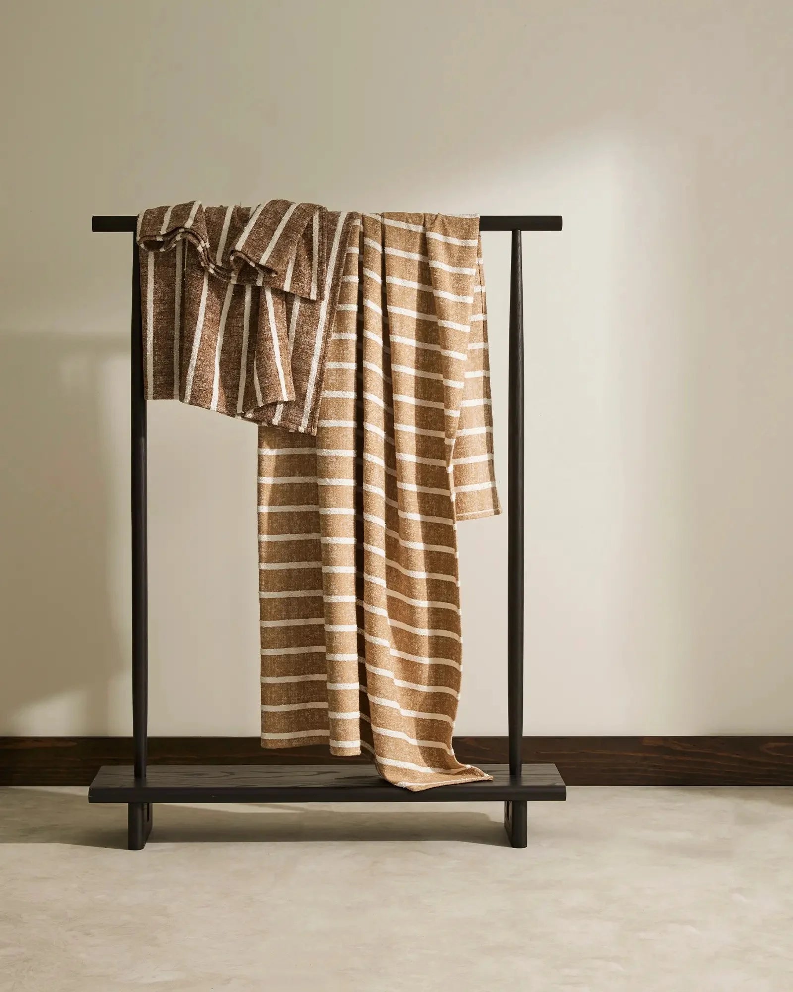 Warm neutral striped throw draped over minimalist black timber rail, showcasing natural linen texture and cream horizontal pattern against soft beige backdrop