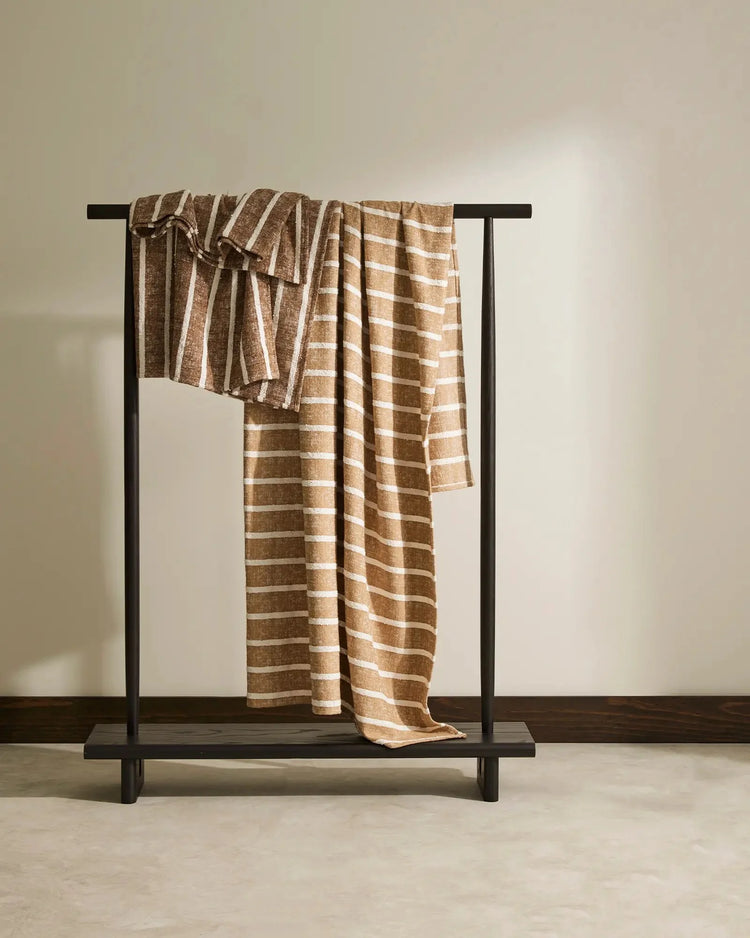 Warm neutral striped throw draped over minimalist black timber rail, showcasing natural linen texture and cream horizontal pattern against soft beige backdrop