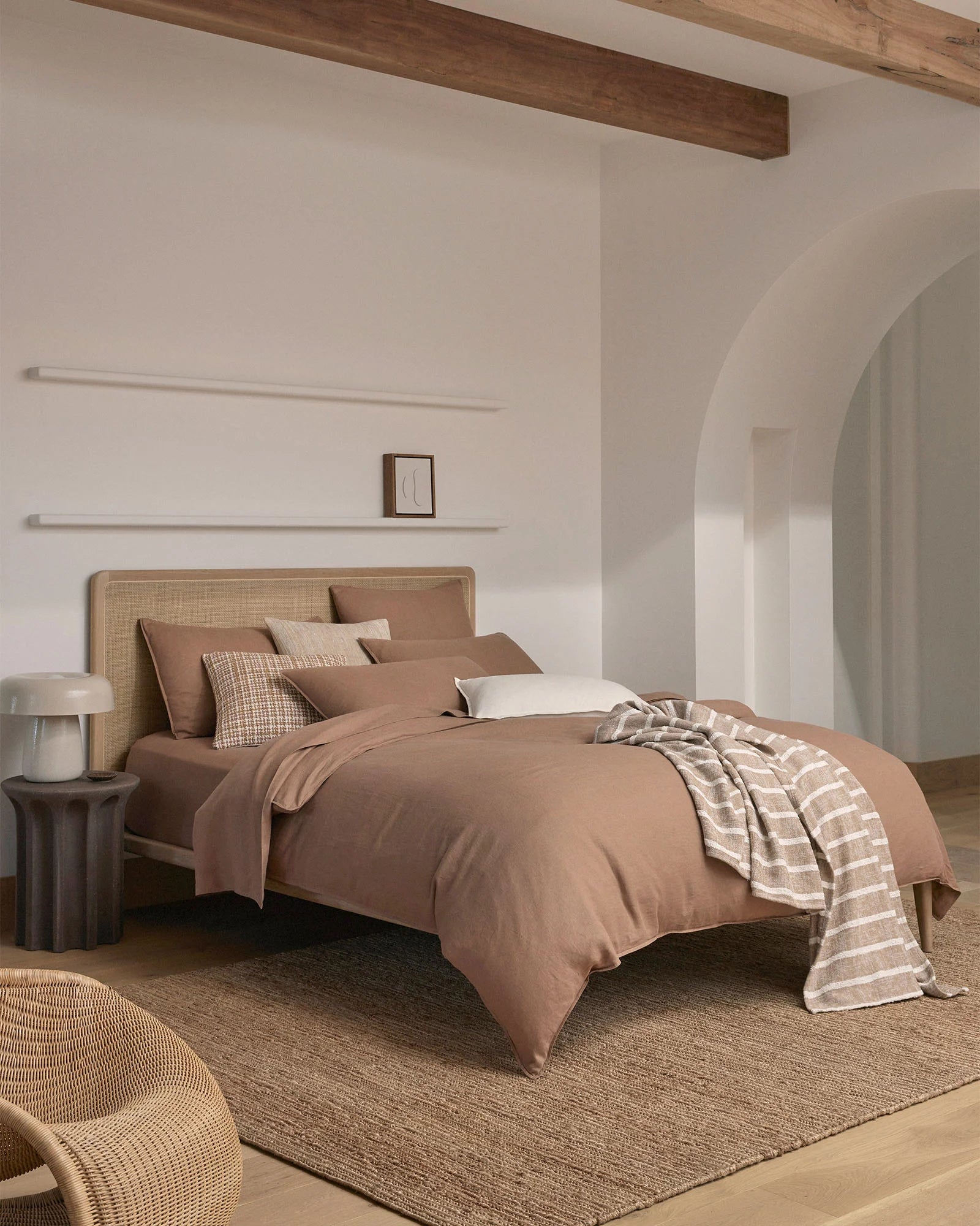 Warm minimalist bedroom with biscuit linen bedding, rattan headboard, and terracotta throw layered over neutral bedding, styled with geometric cushion and natural textures
