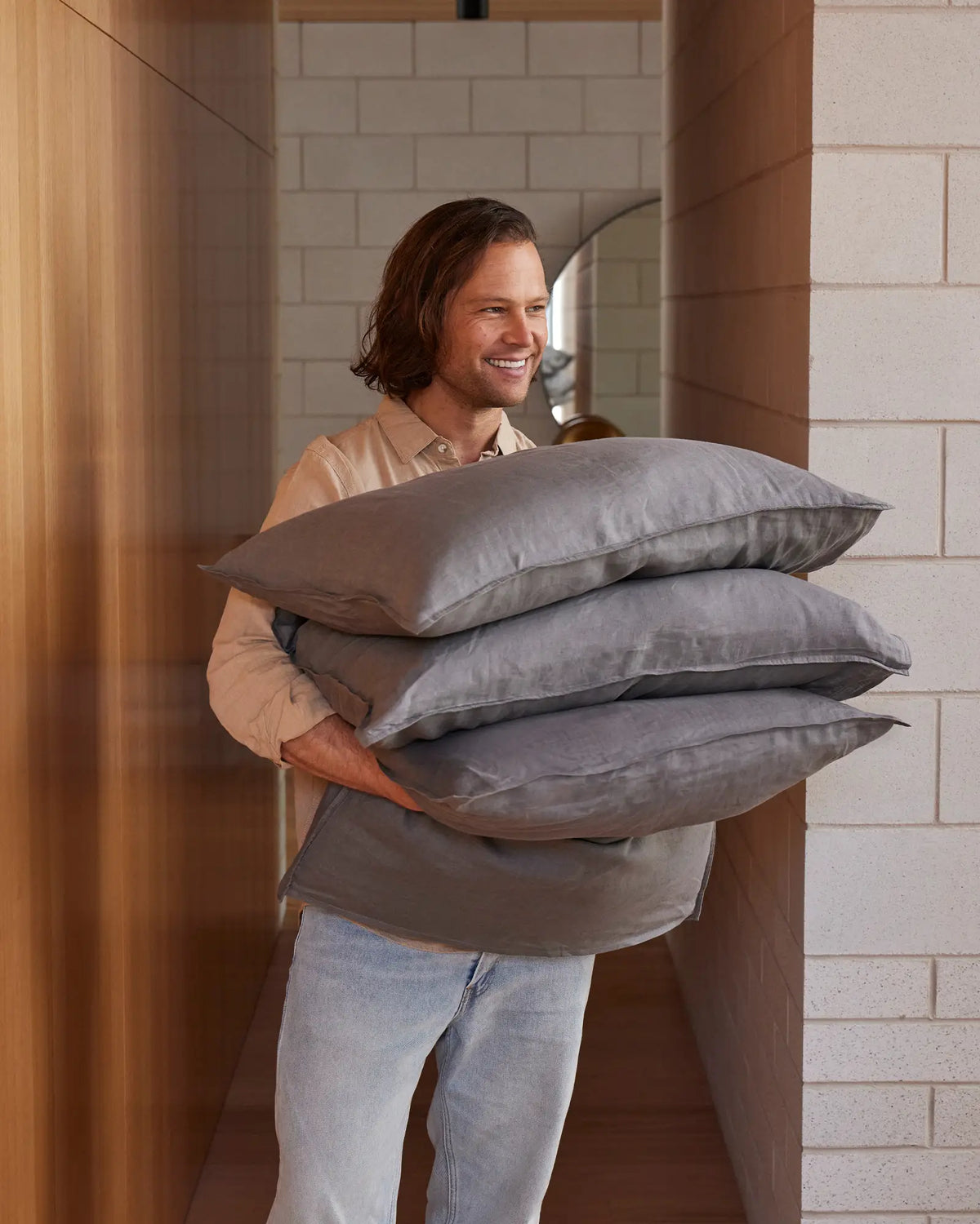 Man holding stack of charcoal linen pillows in warm minimalist hallway, showcasing soft grey bedding textures and earthy colour palette