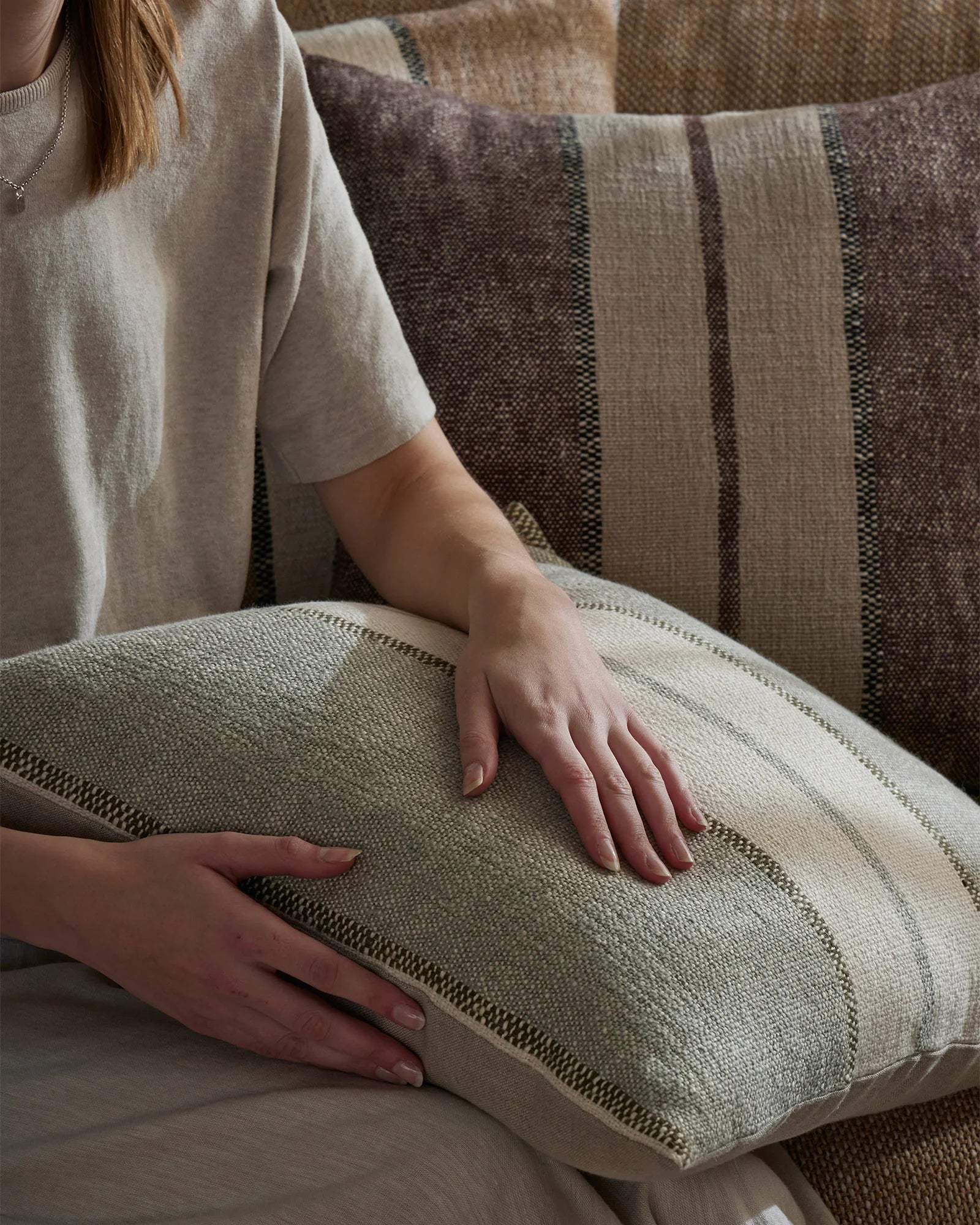Neutral textured cushions with woven striping in sage and taupe tones, styled on a layered sofa arrangement with soft natural lighting and warm tonal palette.