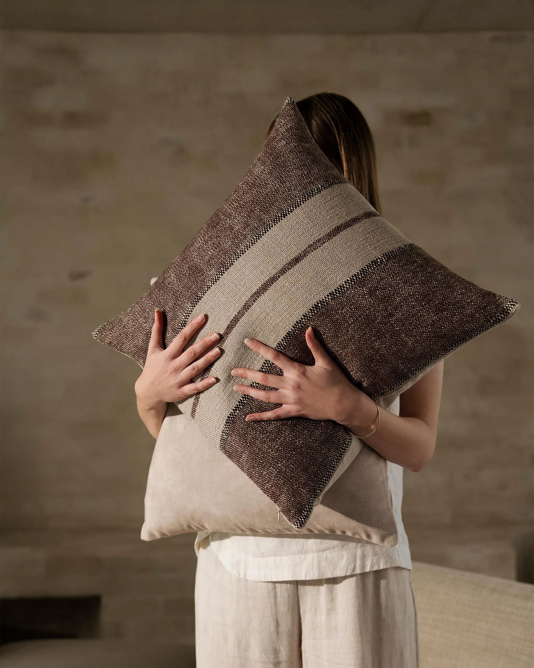 Neutral linen cushion with woven stripe detailing in taupe and charcoal, held against cream textiles in minimalist interior setting