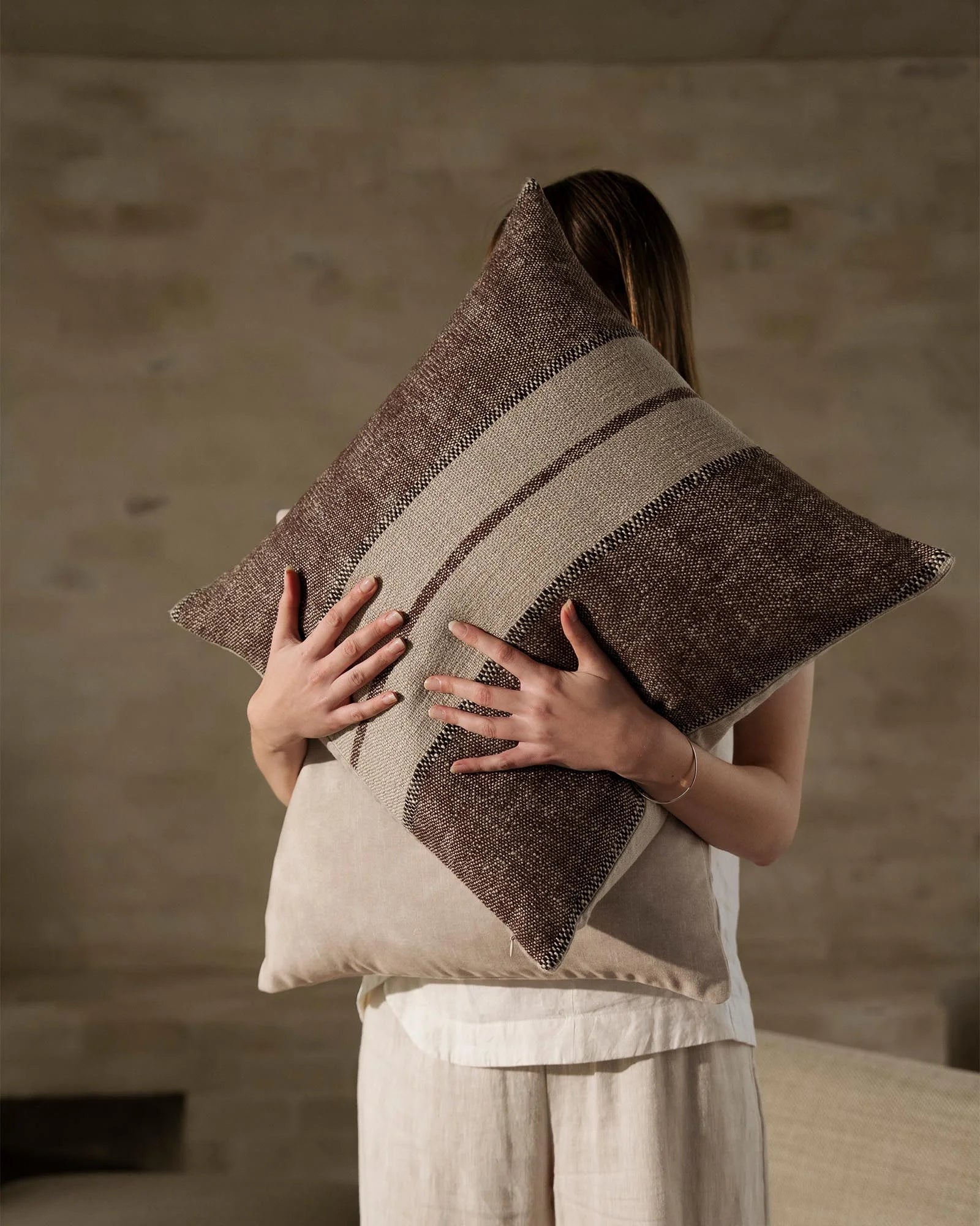 Neutral linen cushion with woven stripe detailing in taupe and charcoal, held against cream textiles in minimalist interior setting
