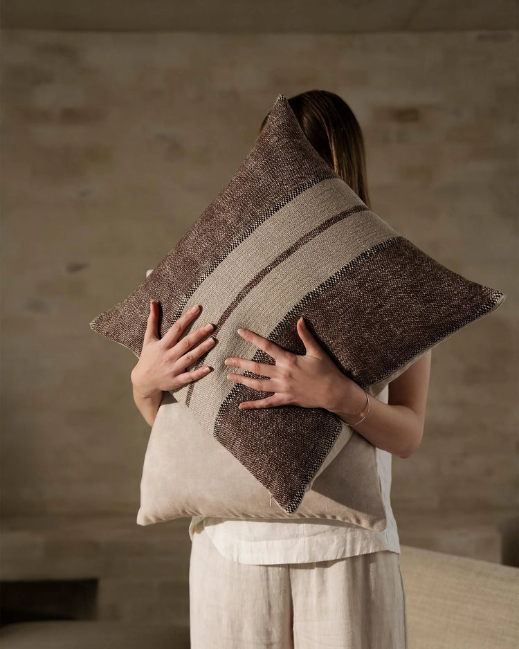 Neutral linen cushion with woven stripe detailing in taupe and charcoal, held against cream textiles in minimalist interior setting