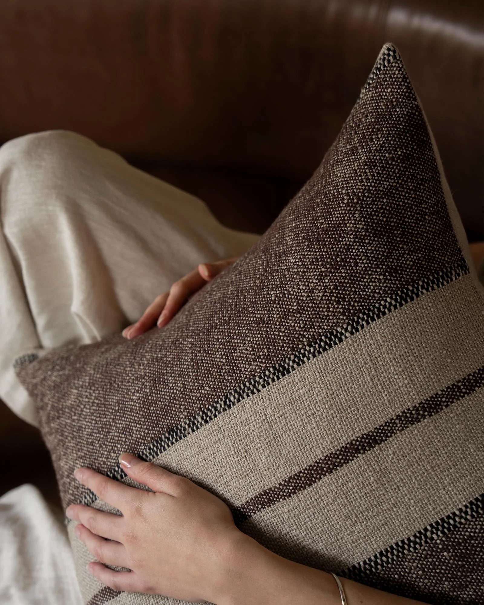 Neutral linen blend cushion with tonal stripe detail draped across cream bedding, earthy brown tones and woven texture creating a warm, minimalist bedroom moment