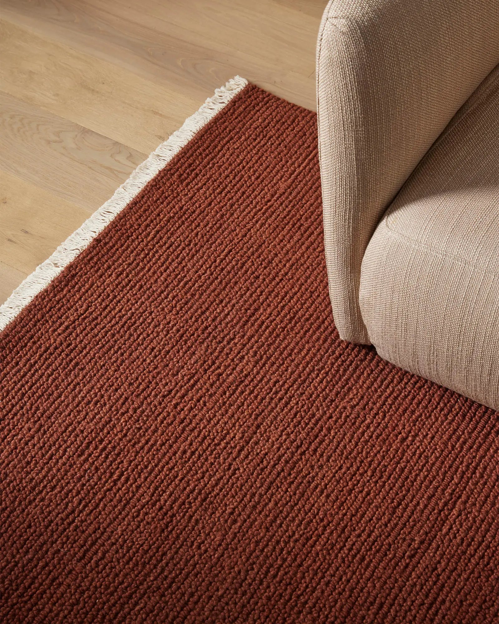 Warm terracotta wool rug with crisp white fringe beside natural beige upholstered furniture on light wood floor, earthy minimalist interior styling