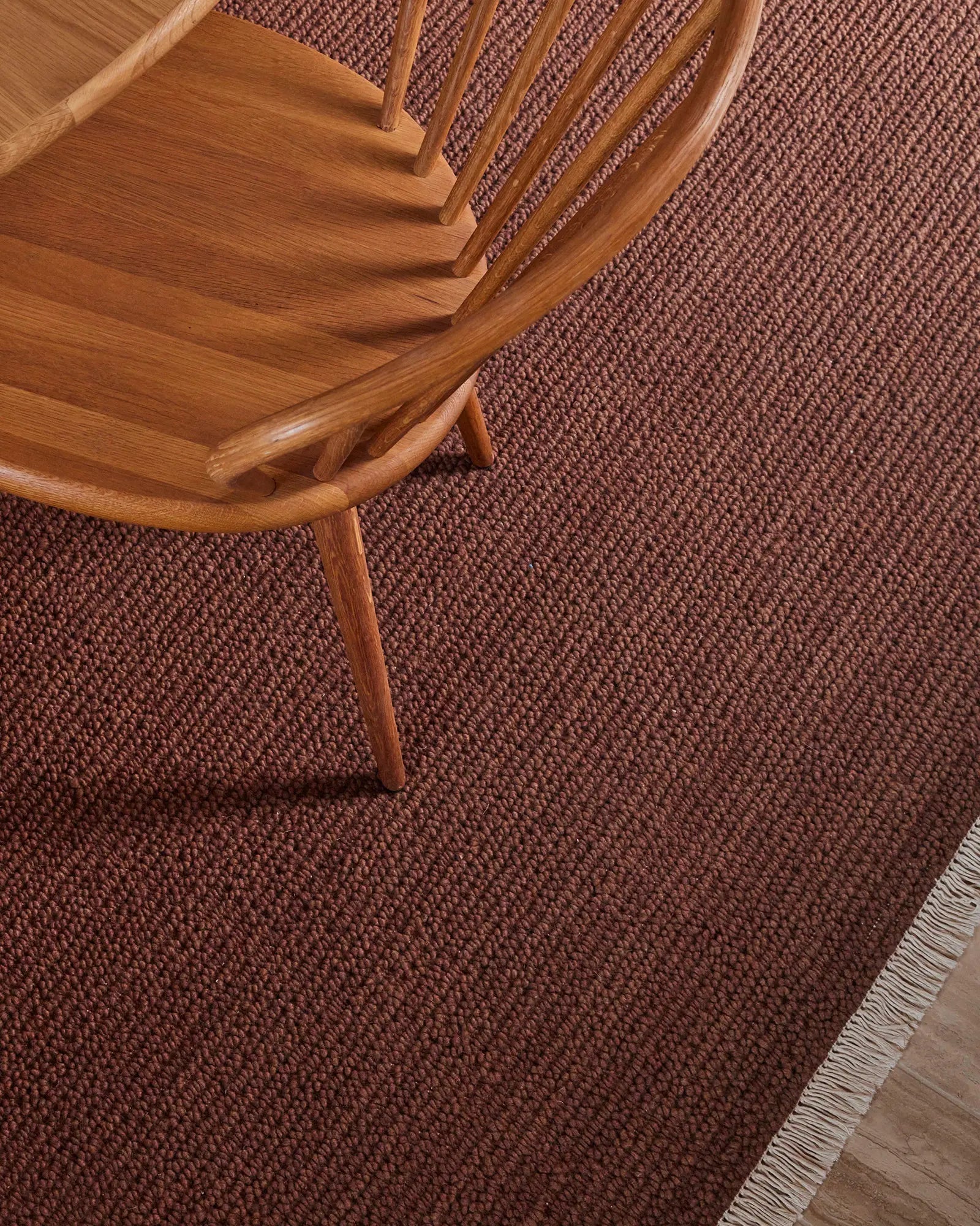 Warm terracotta wool rug with textured weave beneath mid-century wooden chair, styled in earthy, minimalist interior with natural wood tones