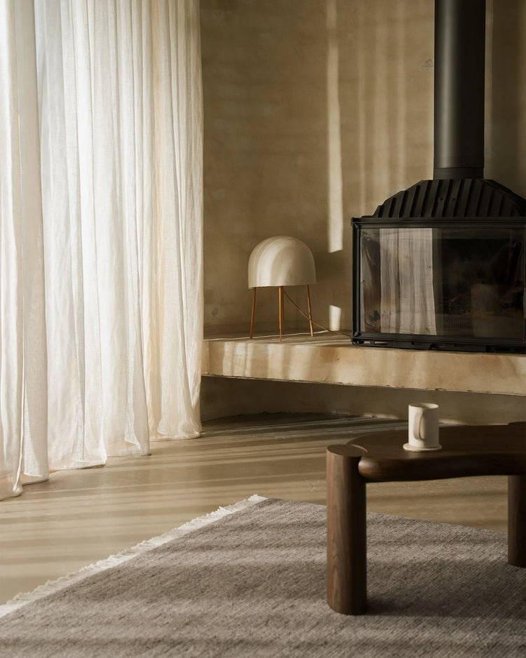 Minimalist living room with warm neutral palette, cream linen curtains filtering soft light, textured ash wool rug anchoring wood furniture and fireplace setting
