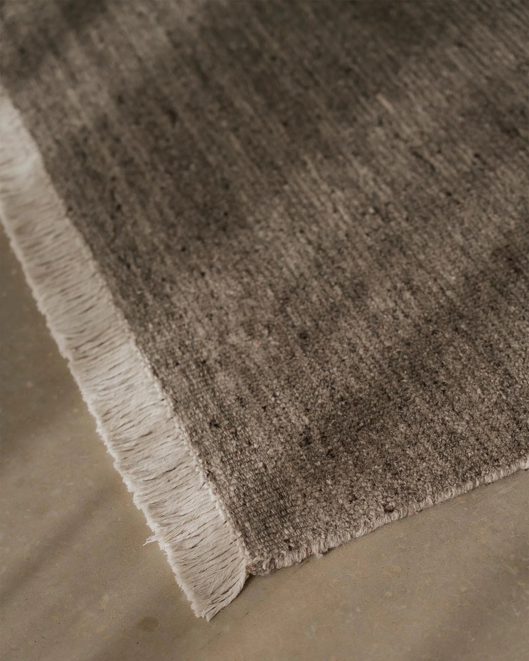 Close-up of Anton Ash rug showing textured wool weave and cream fringe edge on neutral concrete floor