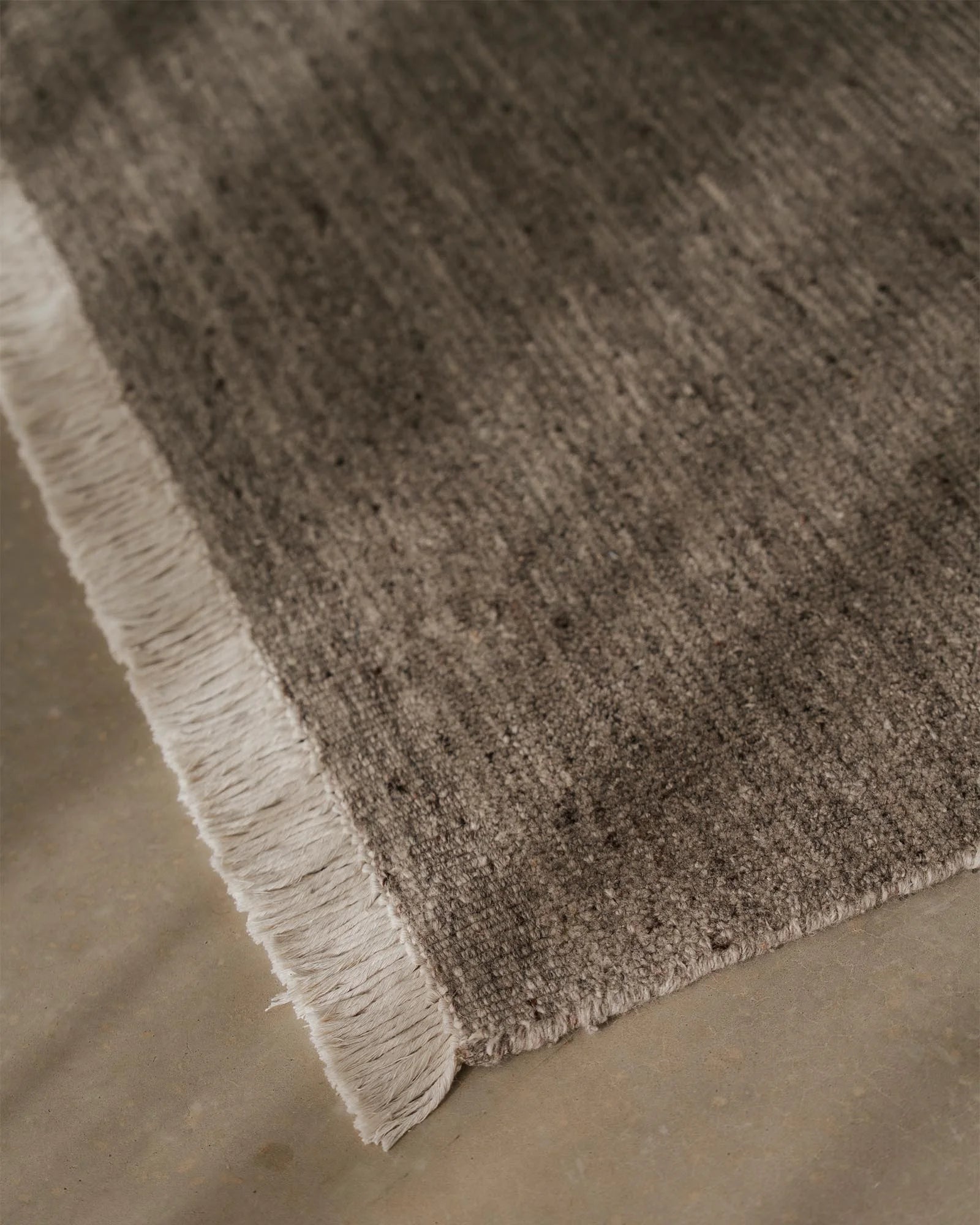 Close-up of Anton Ash rug showing textured wool weave and cream fringe edge on neutral concrete floor