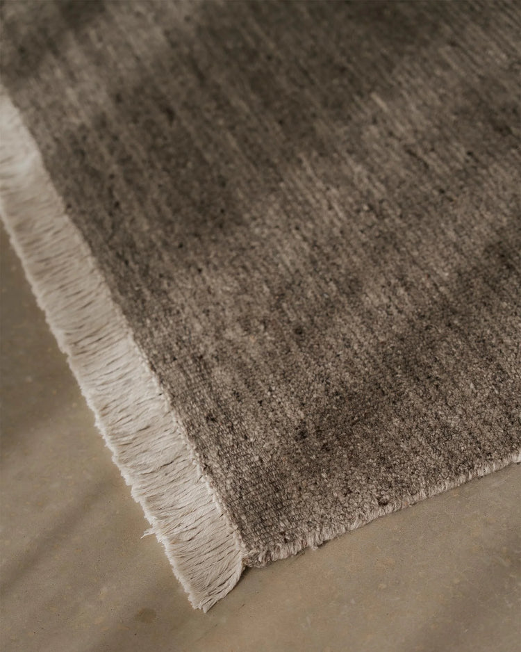 Close-up of Anton Ash rug showing textured wool weave and cream fringe edge on neutral concrete floor