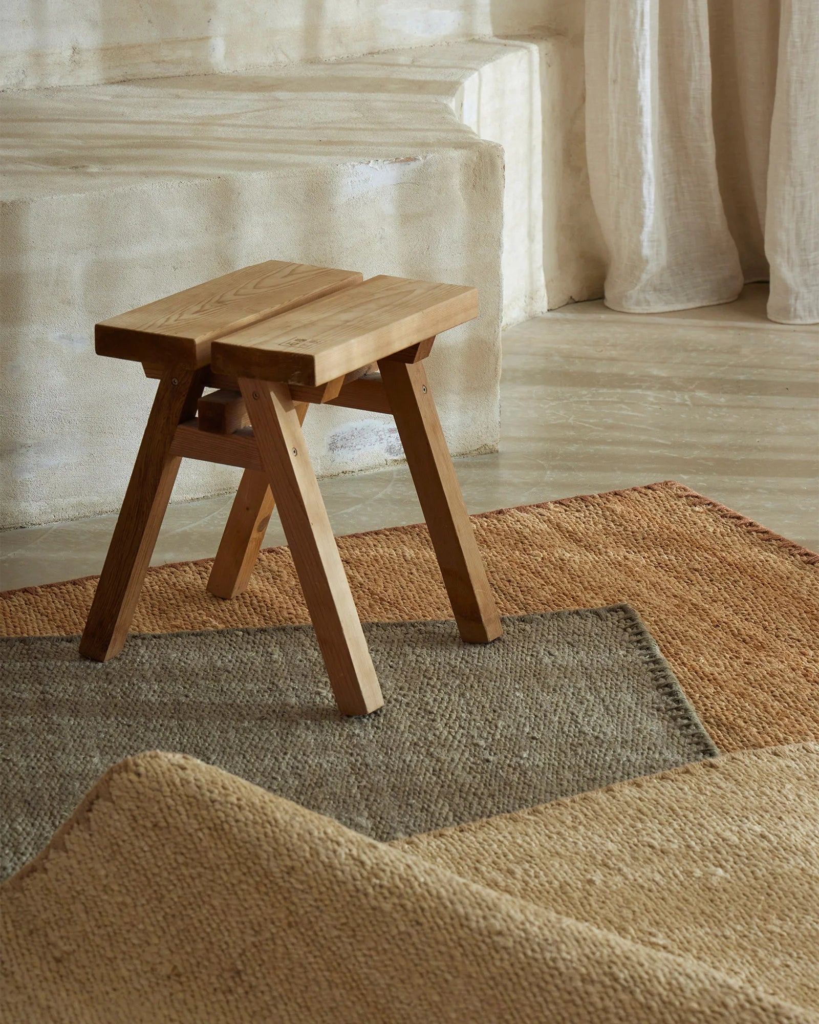 Layered natural fibre rugs in warm oat and cool grey tones anchor a minimalist space with wooden furniture and soft linen curtains, creating earthy neutral styling