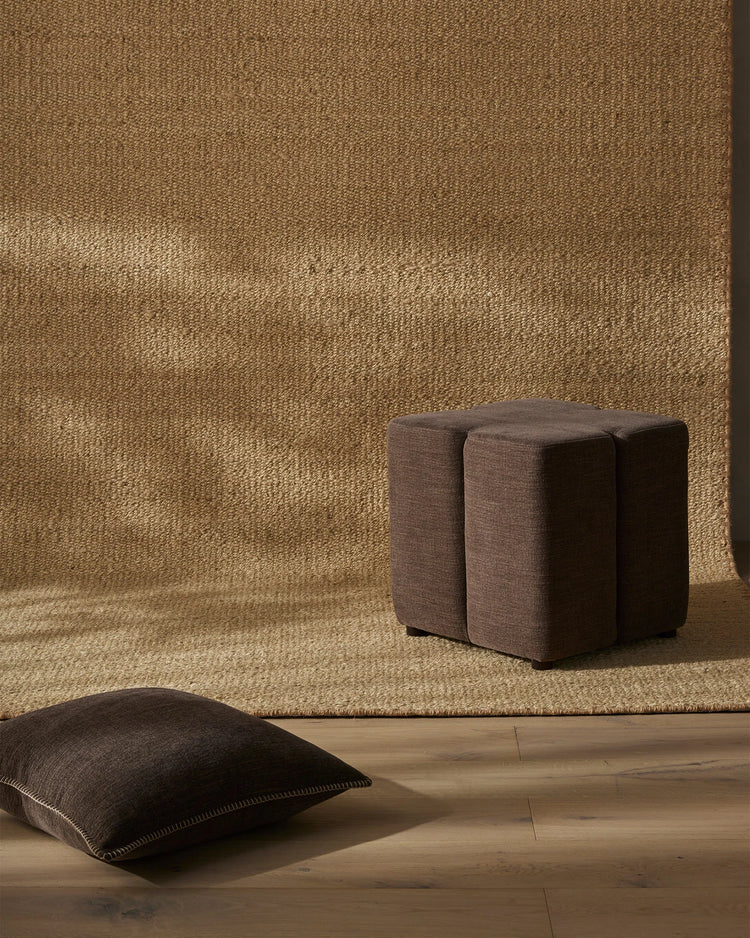 Warm neutral styling with gold textured wallpaper, natural wood flooring, and layered oat-toned rug featuring charcoal linen ottomans and scatter cushions