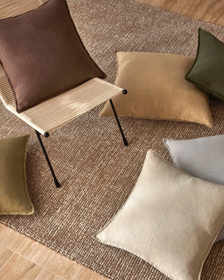 Earthy neutral palette of linen cushions in mocha, olive, cream and taupe arranged on textured jute rug with timber flooring and mid-century chair.