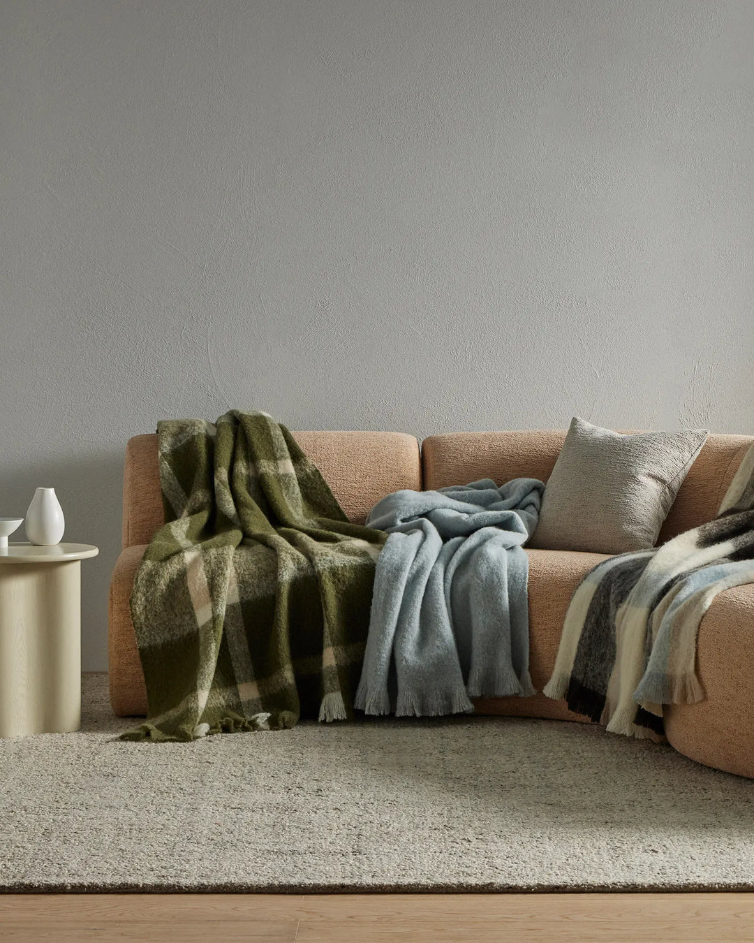 Neutral living room with camel-toned sofa layered with olive plaid, slate blue, and cream wool blend throws; soft textures, earthy palette, minimalist styling.