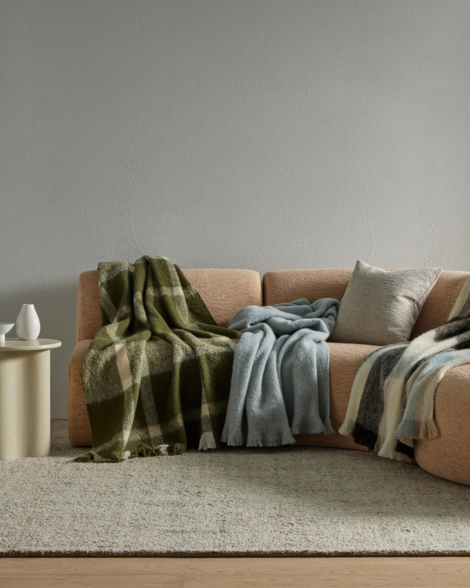 Neutral living room with camel-toned sofa layered with olive plaid, slate blue, and cream wool blend throws; soft textures, earthy palette, minimalist styling.