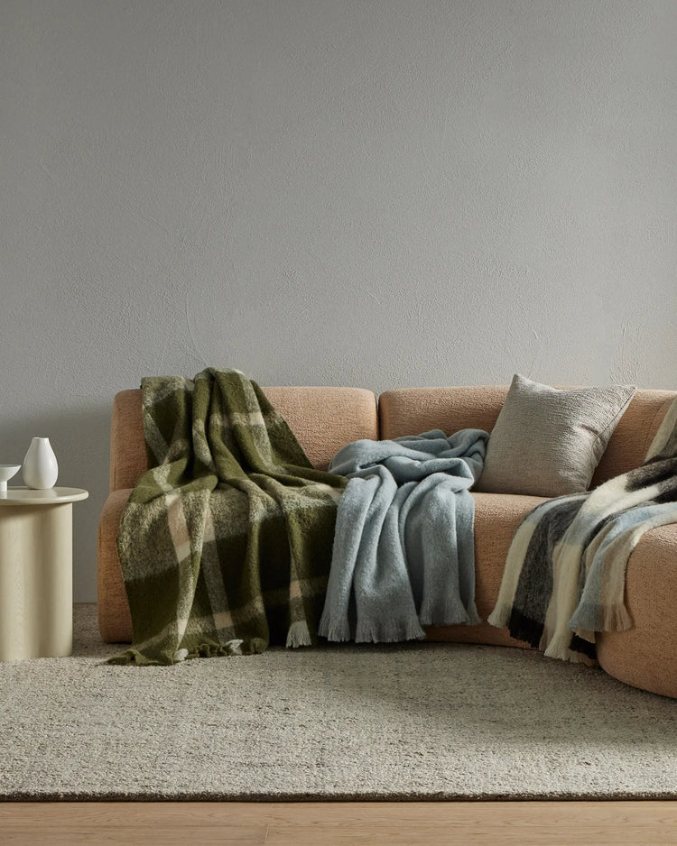 Neutral living room with camel-toned sofa layered with olive plaid, slate blue, and cream wool blend throws; soft textures, earthy palette, minimalist styling.