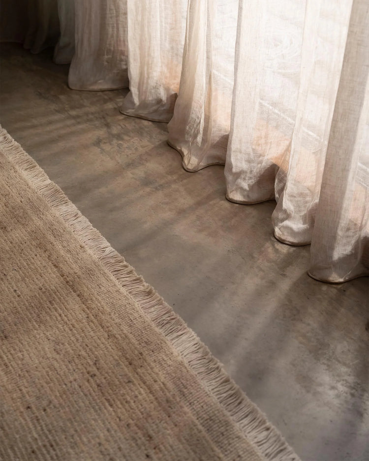 Neutral linen curtains pool on concrete floors beside textured Anton Sand wool rug with fringe trim in warm earthy tones, minimalist industrial aesthetic