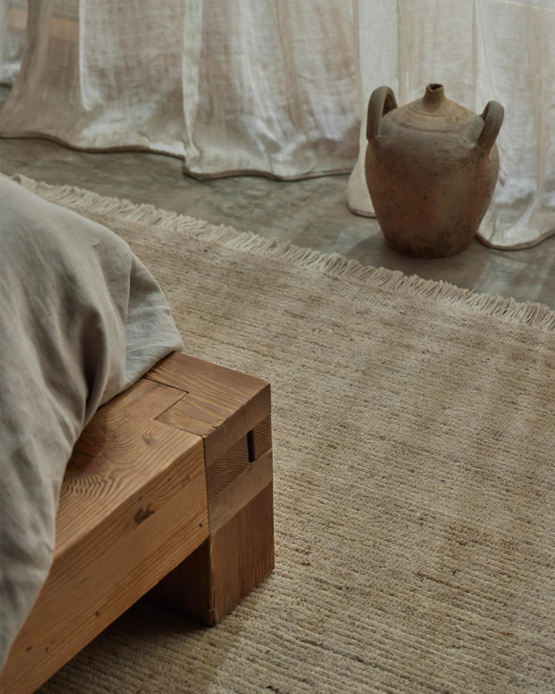 Neutral bedroom styled with creamy linen curtains, natural wood bed frame, and warm terracotta pottery on Anton Sand wool rug in earthy tones