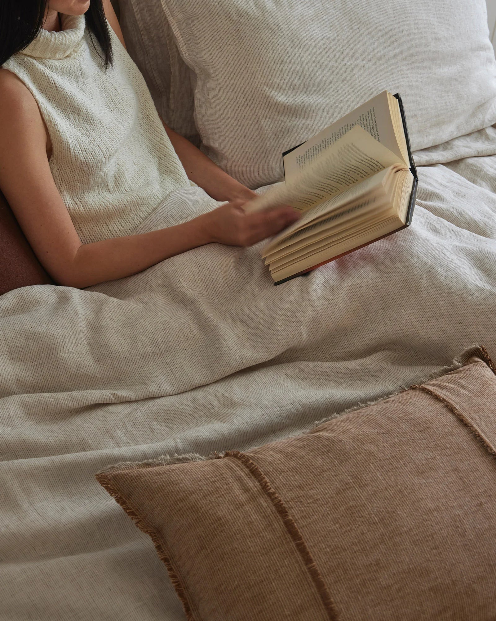 Woman reading on bed layered with neutral linen bedding in cream and taupe tones, featuring textured Sova sheets and Adela rust cushion in natural fibres