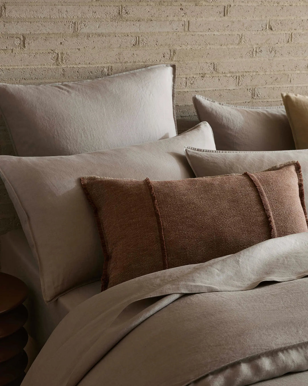 Neutral bed styling with layered linen pillowcases and textured rust-toned cushions against a whitewashed brick wall, earthy minimalist bedroom aesthetic