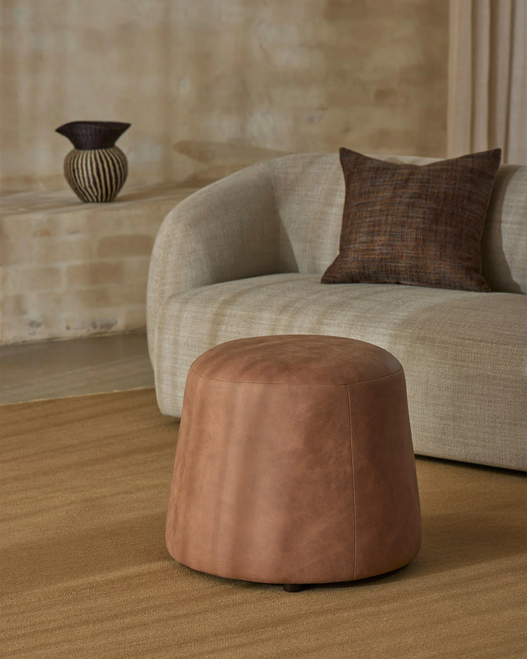 Neutral minimalist living room with cream curved armchair, warm tan wool rug, chocolate linen cushion and cognac leather ottoman on neutral textured backdrop