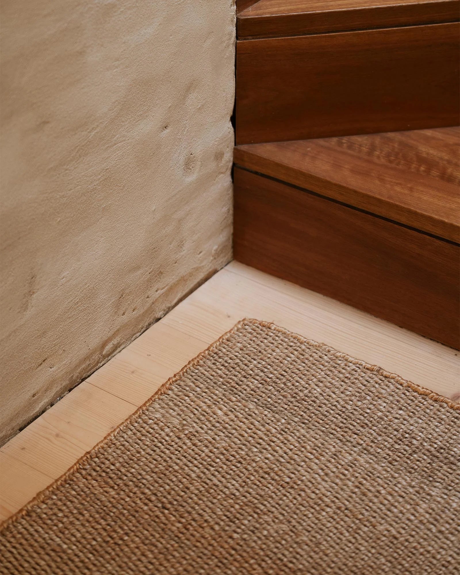 Natural jute doormat layered on pale timber flooring beside warm wood furniture and textured plaster wall, neutral earth-tone entryway styling