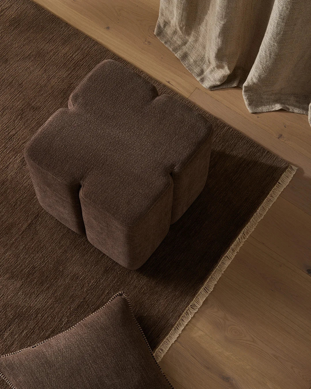 Overhead view of a charcoal linen ottoman styled on a woven rug with natural wood flooring, neutral linen curtains draping beside it, creating a warm minimalist interior.