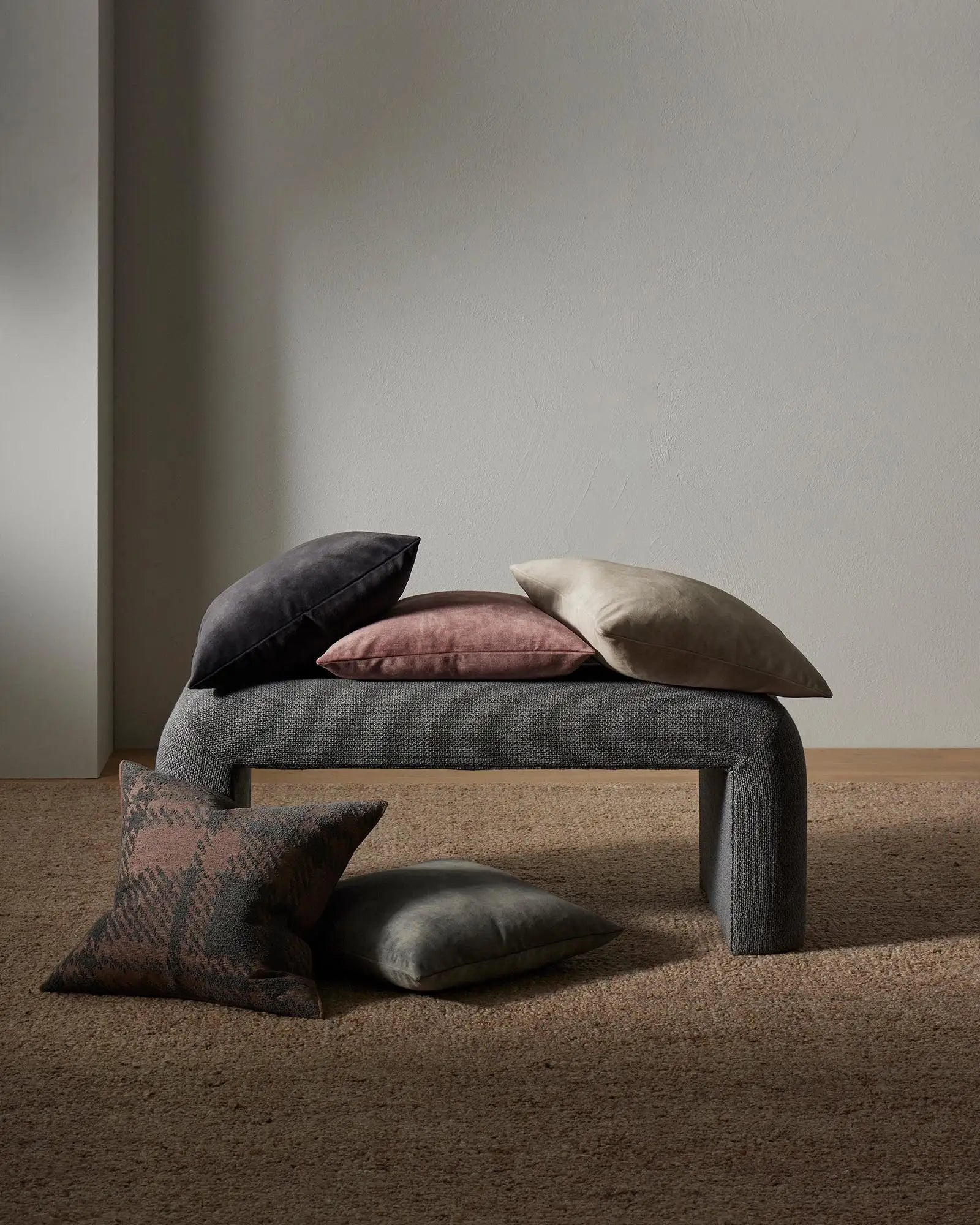 Minimalist living room display with charcoal, mauve and cream cushions arranged on grey upholstered bench, warm linen backdrop and neutral palette styling