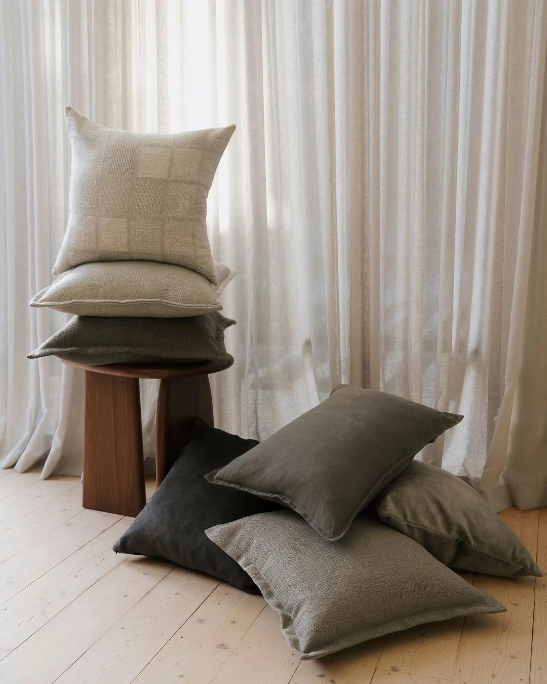 Neutral minimalist interior with stacked cushions in beige, charcoal and grey tones on wooden stool, cream linen drapes, textured fabrics, light timber floors