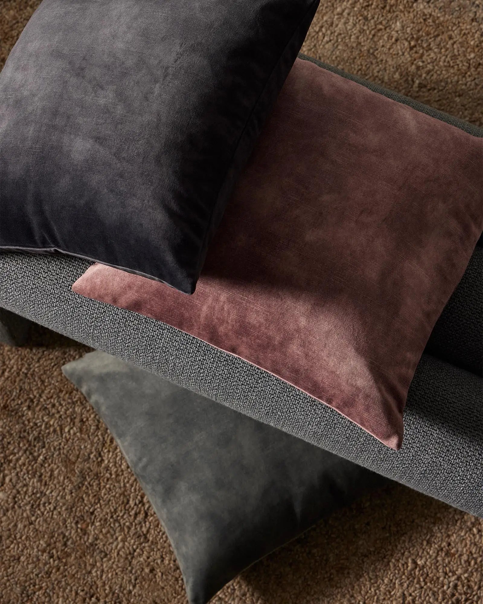 Charcoal and burgundy velvet cushions layered on a grey upholstered sofa with warm taupe textured rug, showcasing moody jewel-tone interior styling
