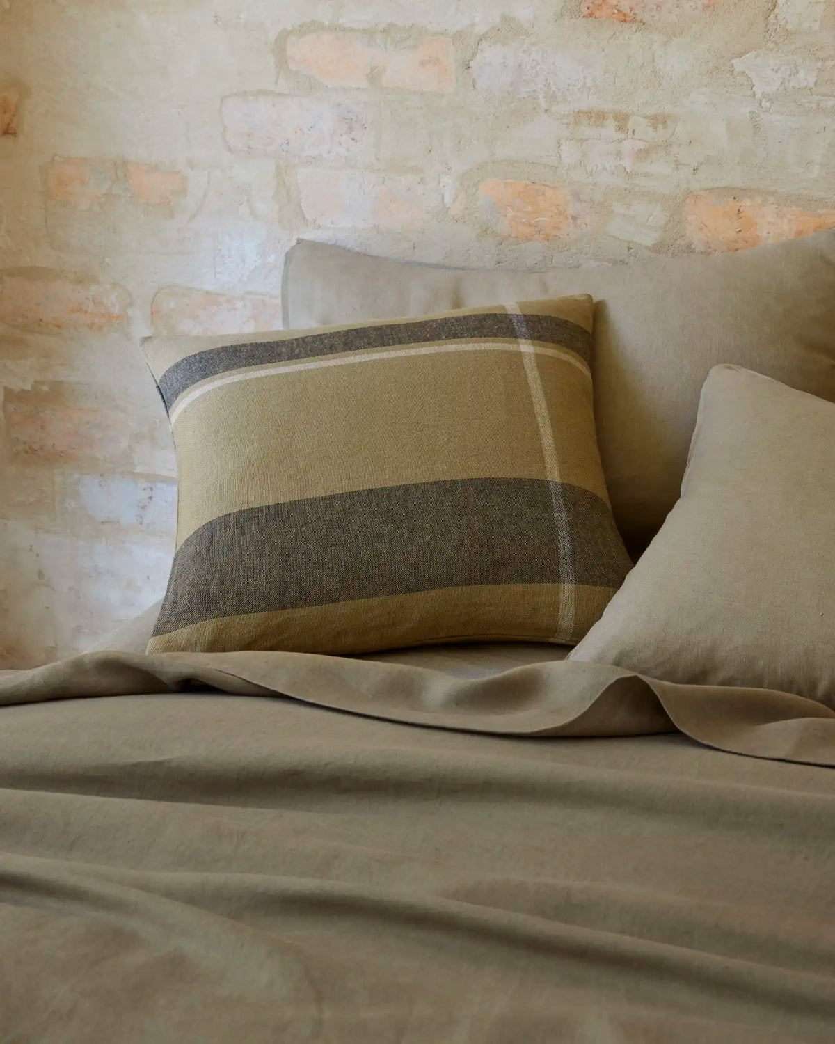 Earthy striped cushion in sage and charcoal on natural linen Ravello bedding, styled against whitewashed brick with soft fabric draping