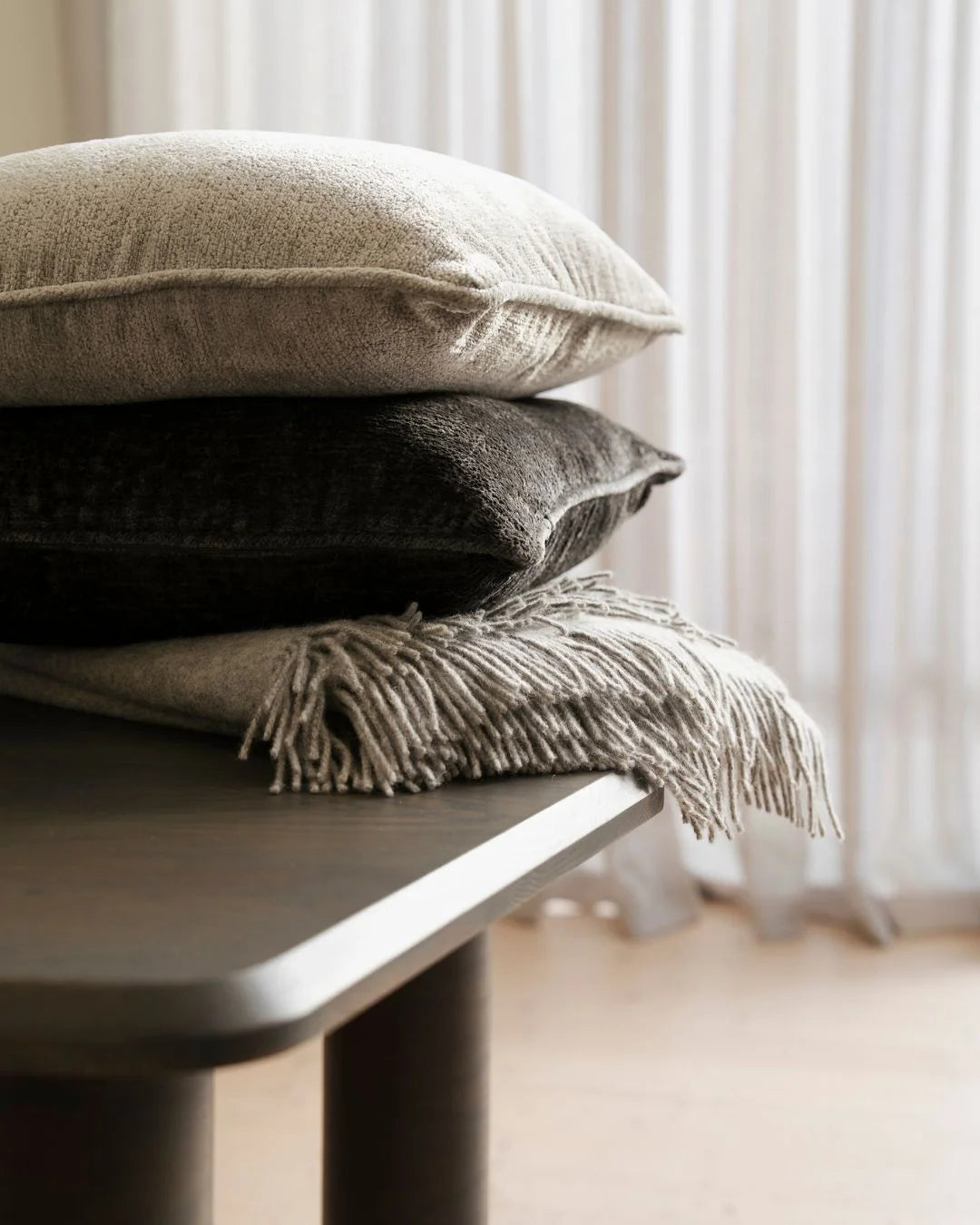 Stacked cushions and throw in neutral tones—charcoal, ash, and warm beige—layered on a modern bench against sheer curtains, showcasing texture contrast and minimalist interior styling