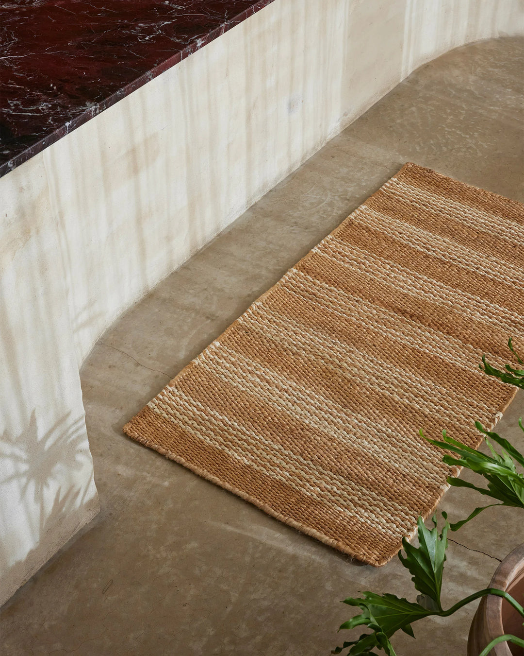 Natural jute door mat layered in neutral minimalist entryway with cream textiles and deep burgundy marble, styled with fresh green foliage